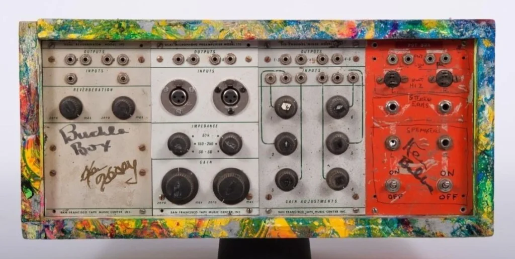 A vintage synthesizer module with colorful, paint-splattered casing, various knobs, switches, and jacks. The words Buchla Box and a signature are written on the left panel.