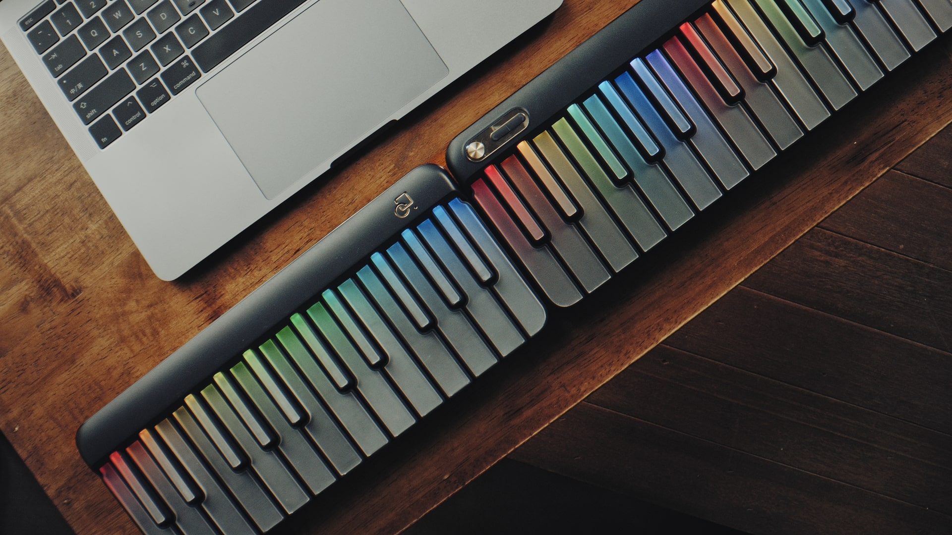 A laptop on a wooden table next to two colorful roll-up piano keyboards with rainbow-colored keys.