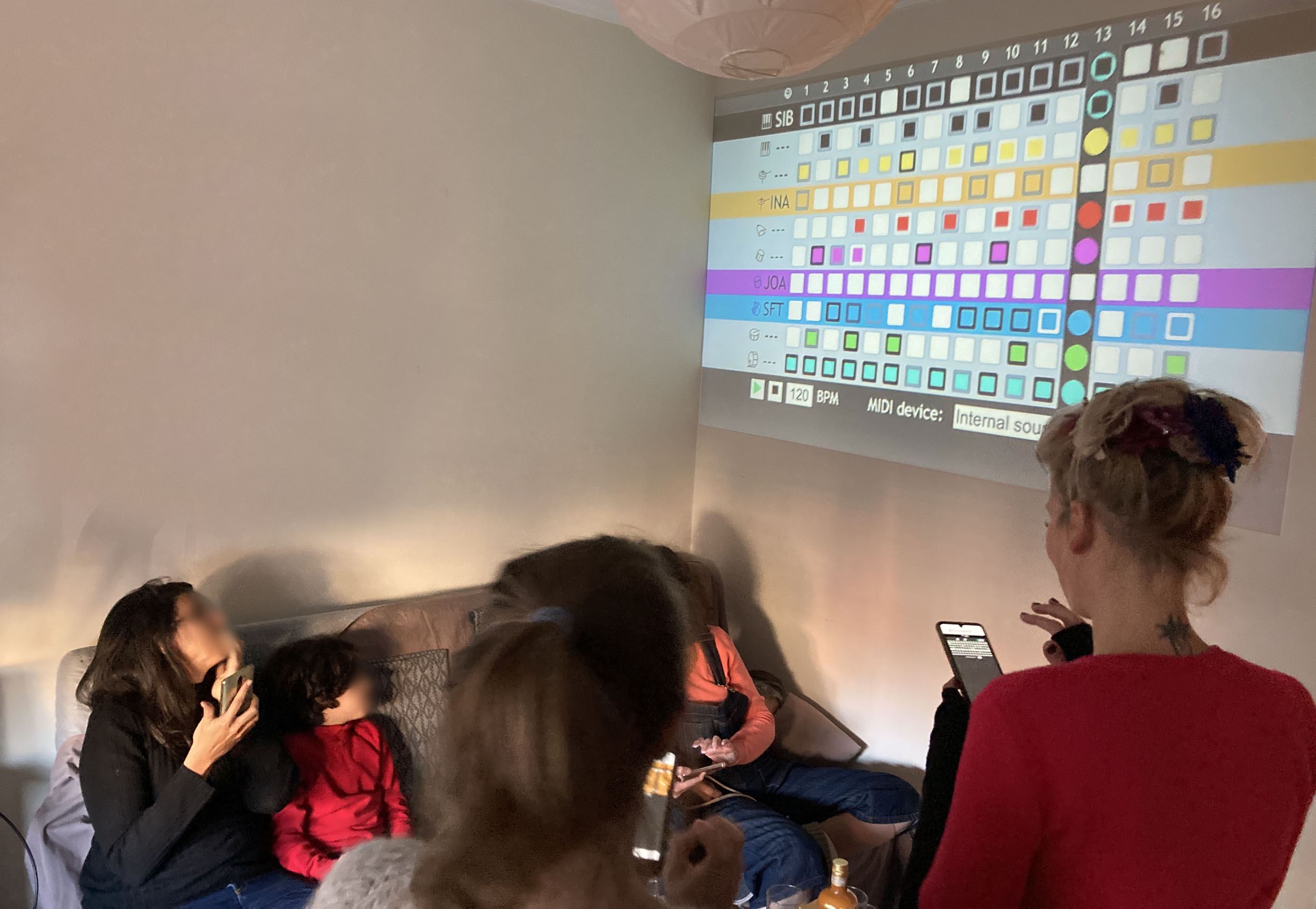 A group of people in a living room look at a wall where a colorful sequencer interface is projected. One person holds a smartphone, and another sits with a child on a couch beneath the projection.