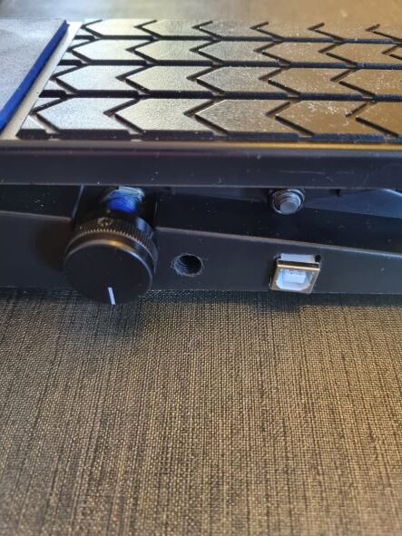 Close-up of a black electronic device with a textured hexagonal pattern, a round control knob, a small button, and a rectangular port, placed on a fabric surface.