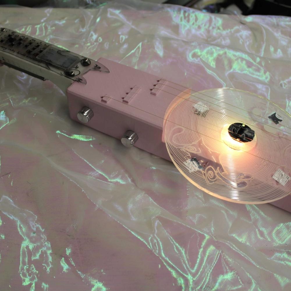 A pink electric guitar with a transparent, glowing circular element near the strings lies on an iridescent, crinkled fabric surface.