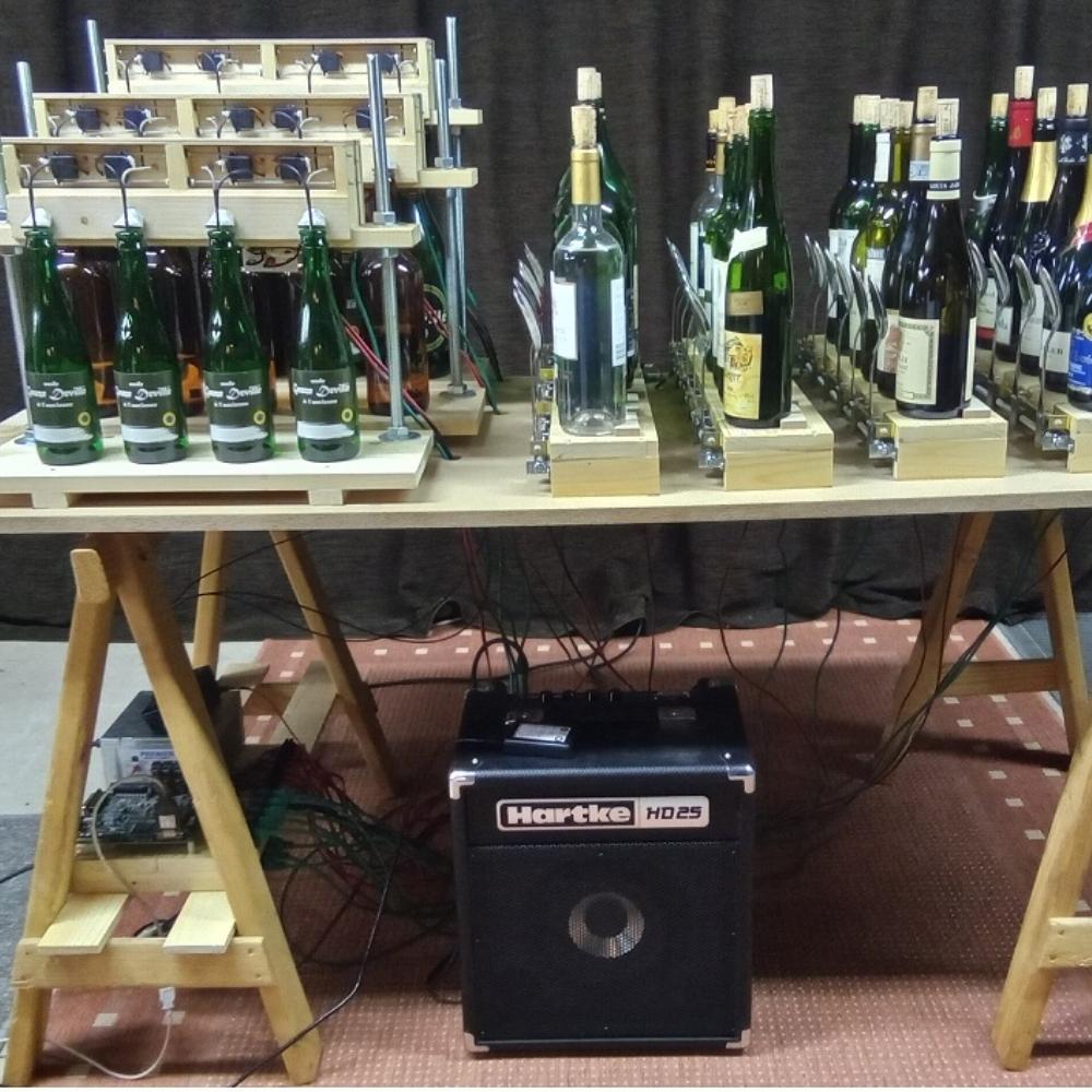 A table holds racks of wine bottles connected to a mechanism with wires and tubes, above a Hartke HD25 amplifier; the setup appears to be a mechanical or musical device involving bottles.