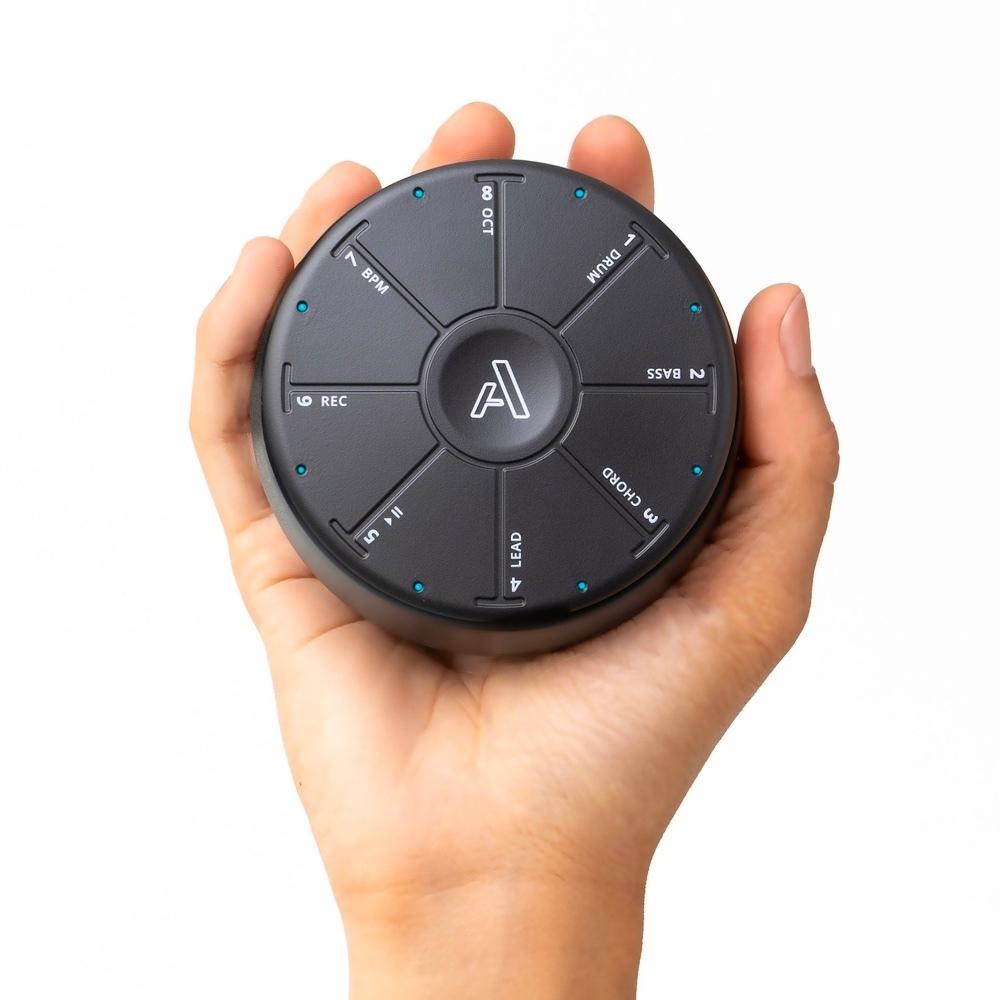 A hand holding a round, black electronic device with buttons labeled with functions such as REC, BASS, and KIT. The device has a central logo and small blue indicator lights around its edge.