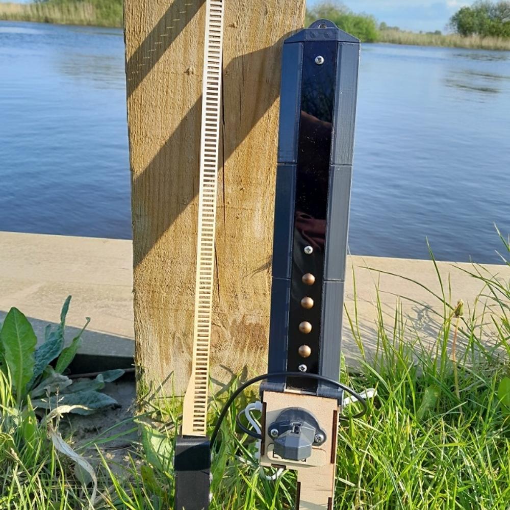 A close-up of a vertical measuring gauge and a black electronic device attached to a wooden post by the water’s edge, surrounded by grass and plants, with a river and trees in the background.