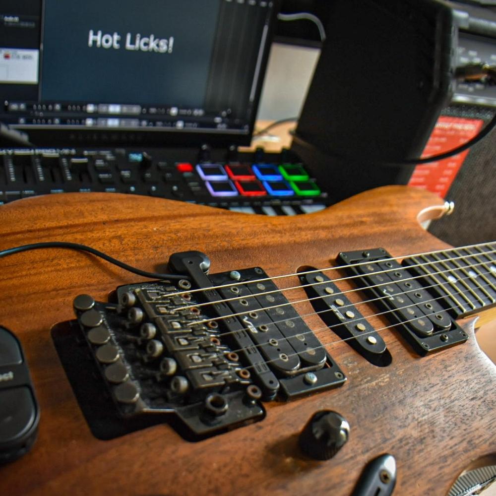 A close-up of an electric guitar with a wooden finish, lying in front of audio equipment and a computer screen displaying the words Hot Licks! in a music production setup.
