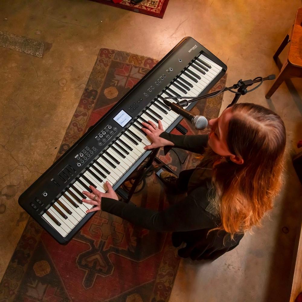 A woman with long hair plays an electric keyboard and sings into a microphone, viewed from above. She is seated on a patterned rug in a warmly lit room.