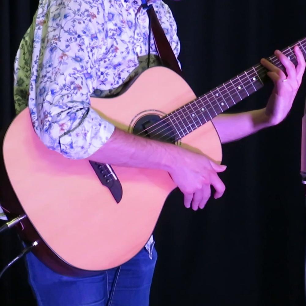 A person wearing a floral-patterned shirt and jeans plays an acoustic guitar on stage, with their hands positioned on the fretboard and strings. The background is dark, highlighting the musician.