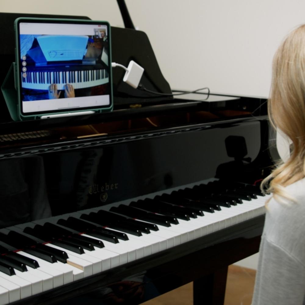 A person with blonde hair sits at a black grand piano, watching an online piano lesson displayed on a tablet propped up on the piano, showing piano keys and hands playing.