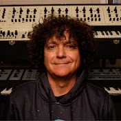 A person with curly dark hair and a slight smile wears a dark hoodie, standing in front of stacked synthesizers with various knobs and keys.