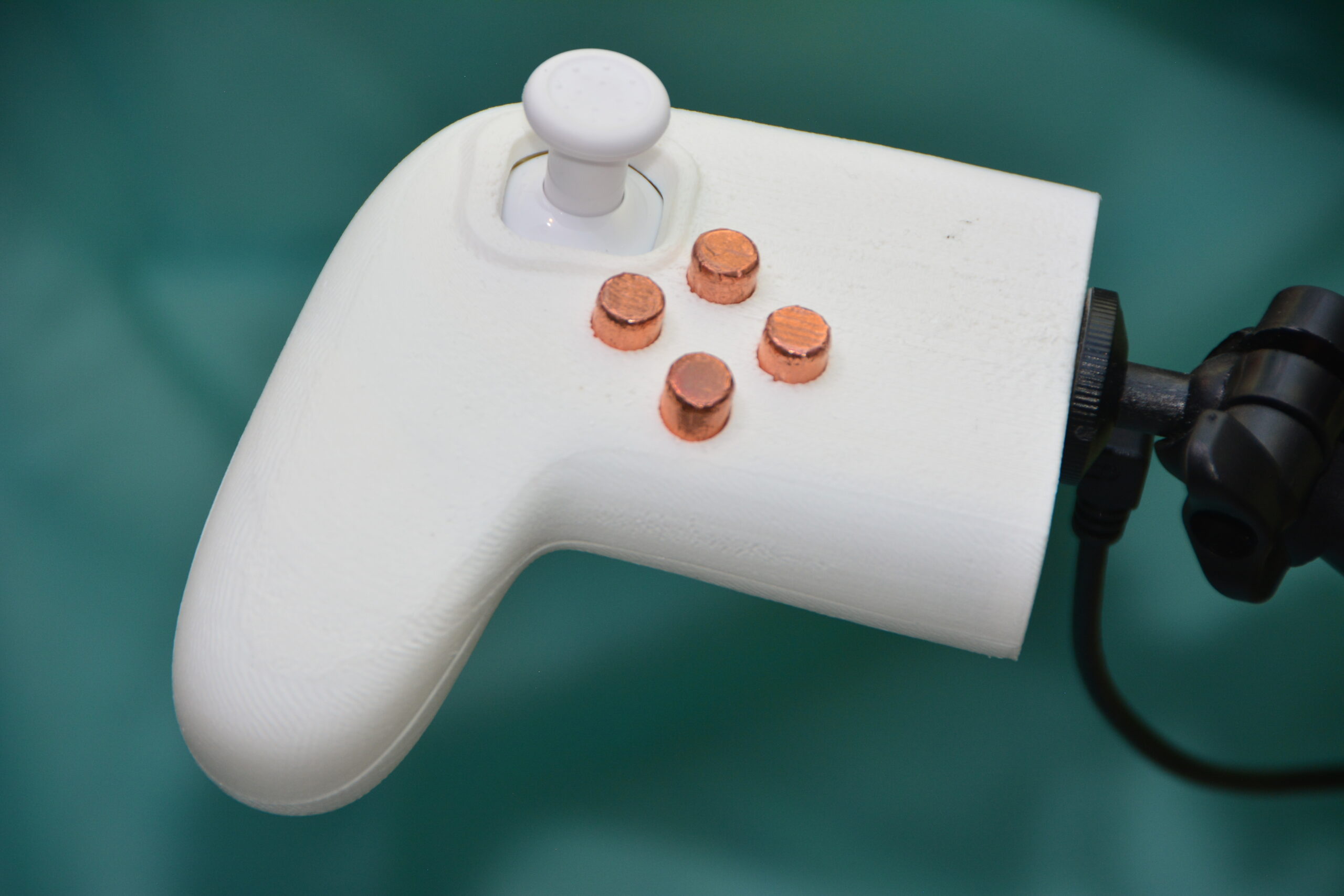 A close-up of a white, 3D-printed game controller with copper-colored circular buttons and a white joystick, set against a teal background.