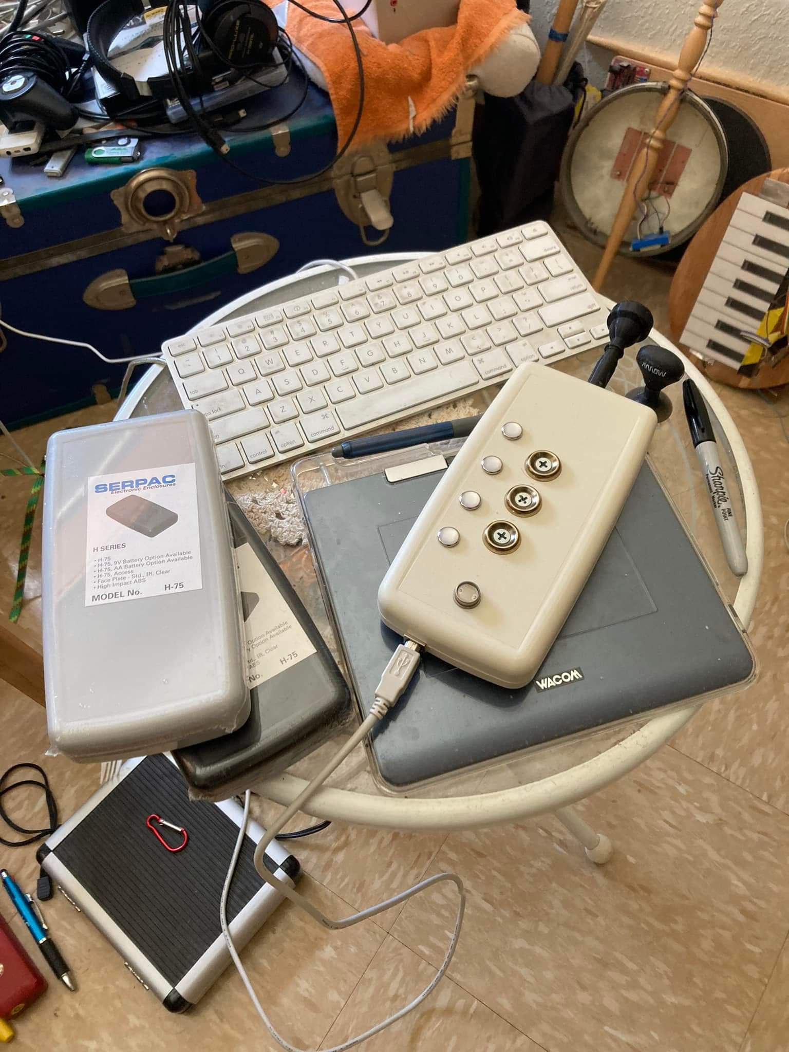 A cluttered round table holds a computer keyboard, two Wacom tablets, a white device with buttons and a cable, two BERPAC cassette tapes, earphones, a marker, and various other electronics and cables.
