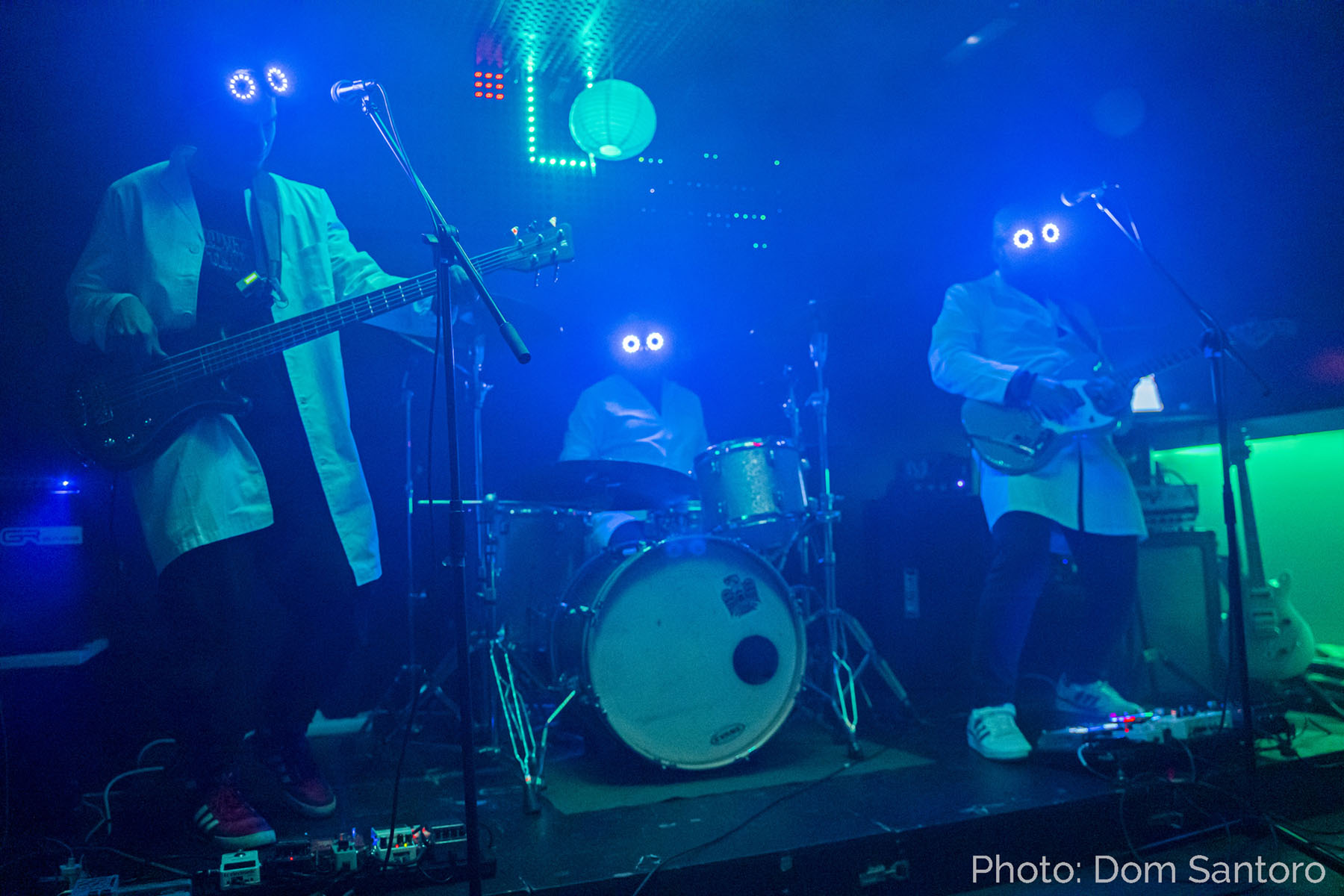 Three musicians wearing lab coats and glowing goggles perform on stage with blue and green lighting, playing bass, drums, and guitar. Photo credit: Dom Santoro.