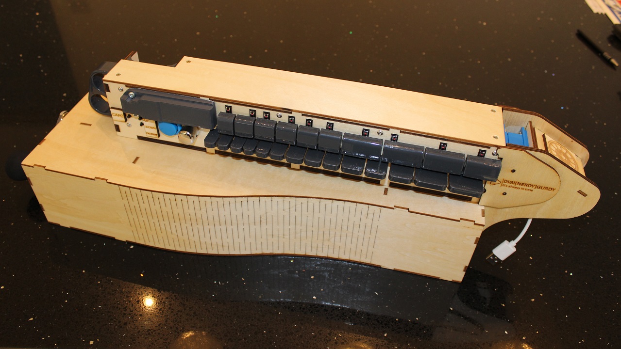 A wooden mechanical device with black keys, resembling a typewriter or musical instrument, sits on a glossy black surface. It has labeled keys and visible internal components.