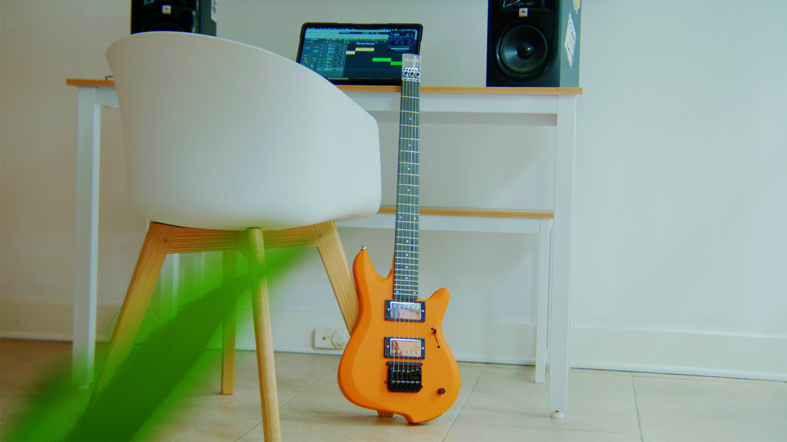 A bright orange electric guitar rests against a white desk with a tablet showing music software, flanked by two speakers. A white chair and a green plant are partially visible in the foreground.