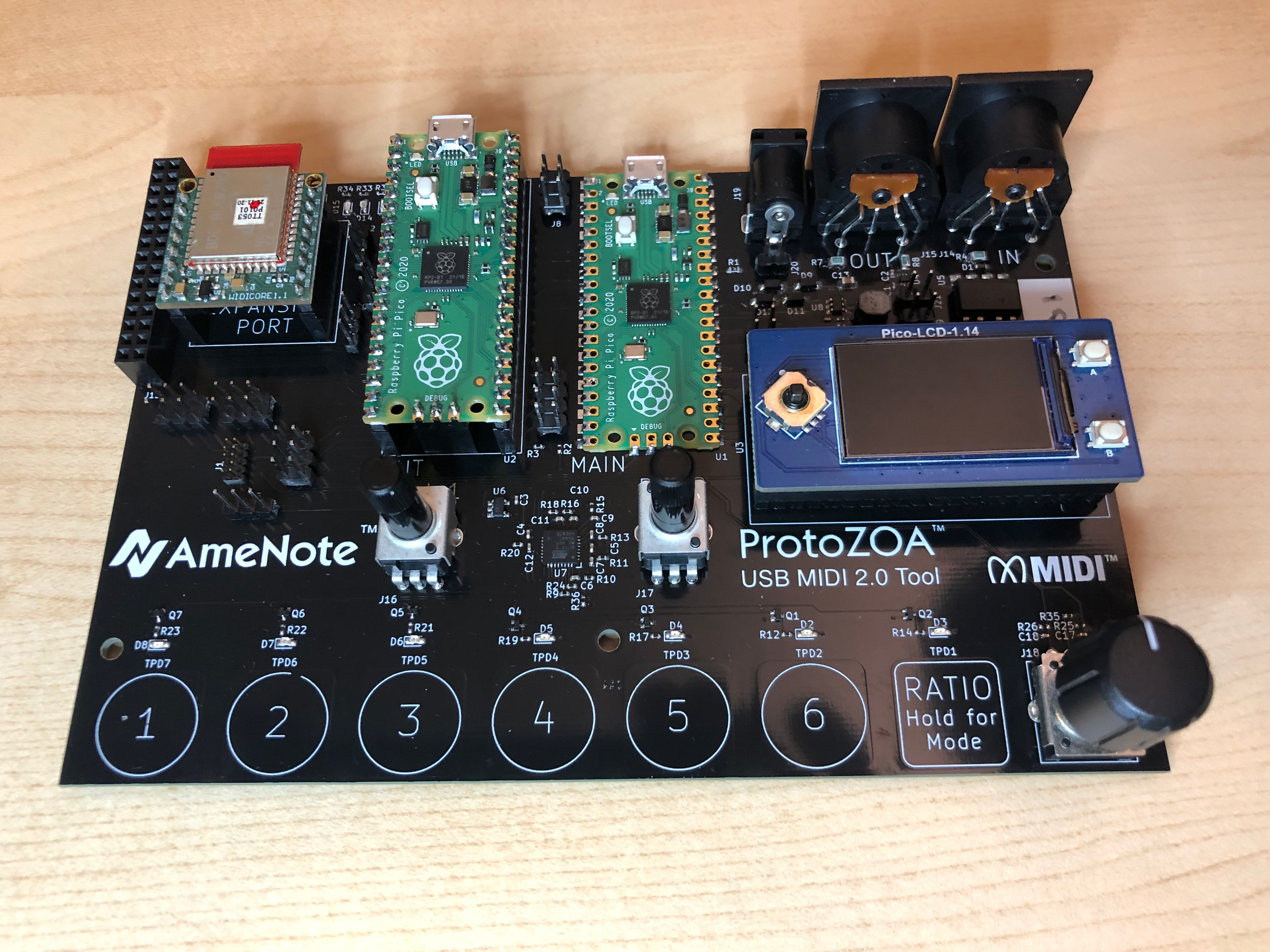 A ProtoZOA USB MIDI 2.0 Tool circuit board by AmeNote, featuring microcontroller modules, an OLED display, various ports, buttons, and a rotary knob, all set on a light wooden surface.