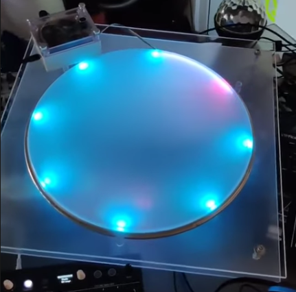 A round, frosted disc on a transparent square base is lit by blue and pink LED lights around its edge, with electronic components and cables visible nearby.