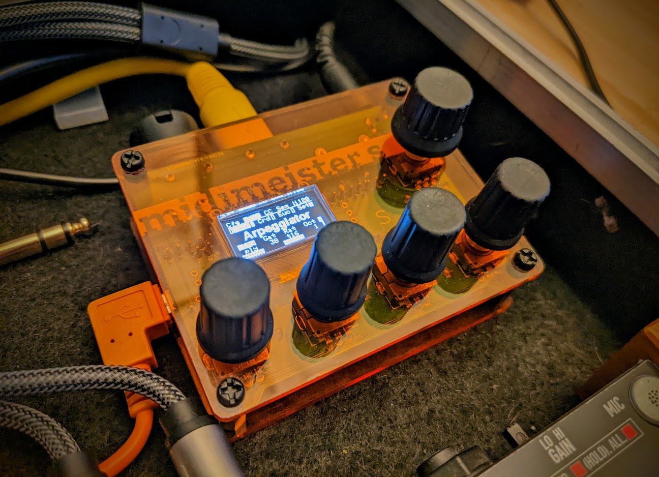 A small orange MIDI controller with five black knobs and a small digital screen displaying “Arpeggiator on/off.” Various cables are plugged in around the device, which sits on a black surface.