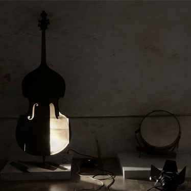 A double bass and a tambourine are displayed on separate platforms in a dimly lit room. Light shines through the double bass, casting shadows, with a spotlight and power cords nearby.