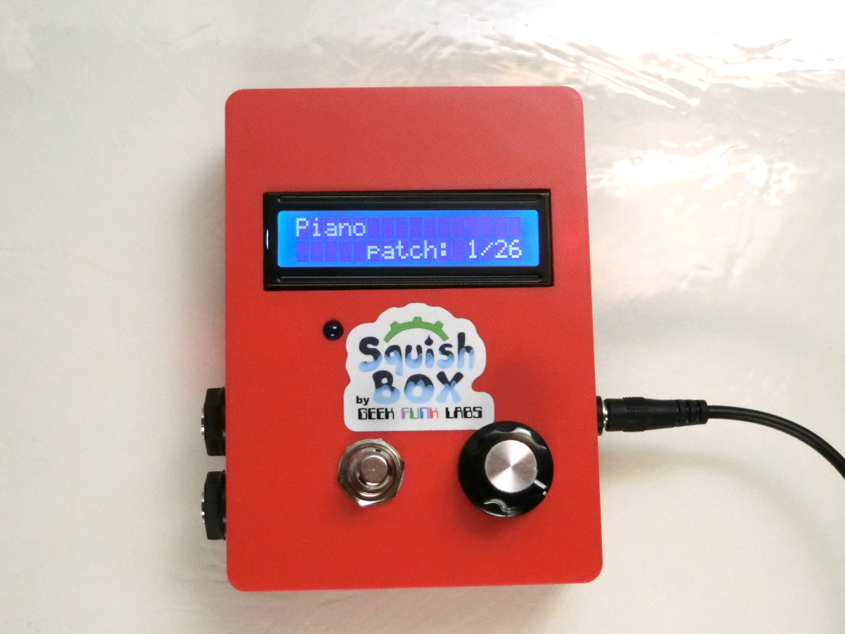 A red electronic device labeled “Squish Box” displays “Piano Patch: 1/26” on its screen. It has two knobs, a button, and a cable plugged in on the right side.