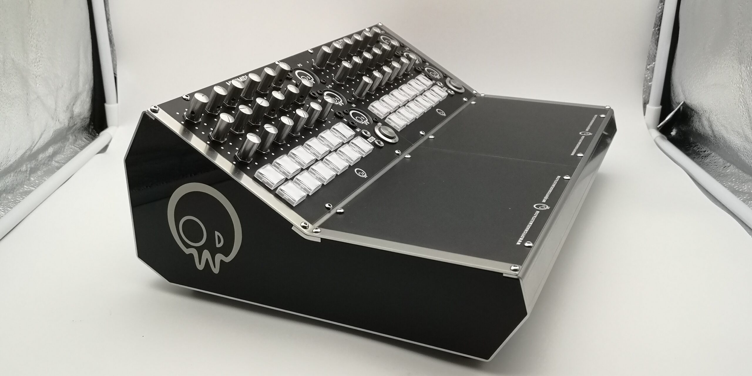 A black audio mixing console with multiple knobs, buttons, and sliders sits on a white surface. The side features a stylized skull logo. The background is neutral and softly lit.