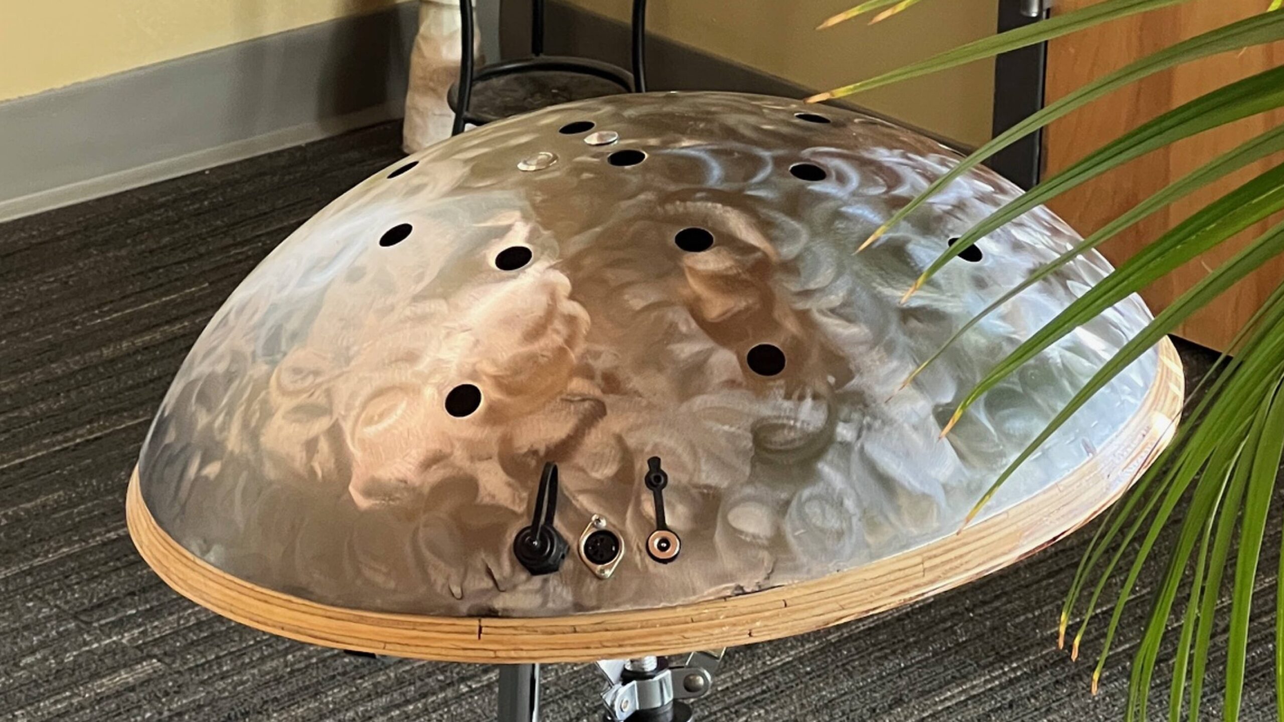 A metallic handpan drum with multiple circular holes sits on a stand, featuring electronic input/output jacks and switches on its side. A green plant leaf is visible in the foreground.