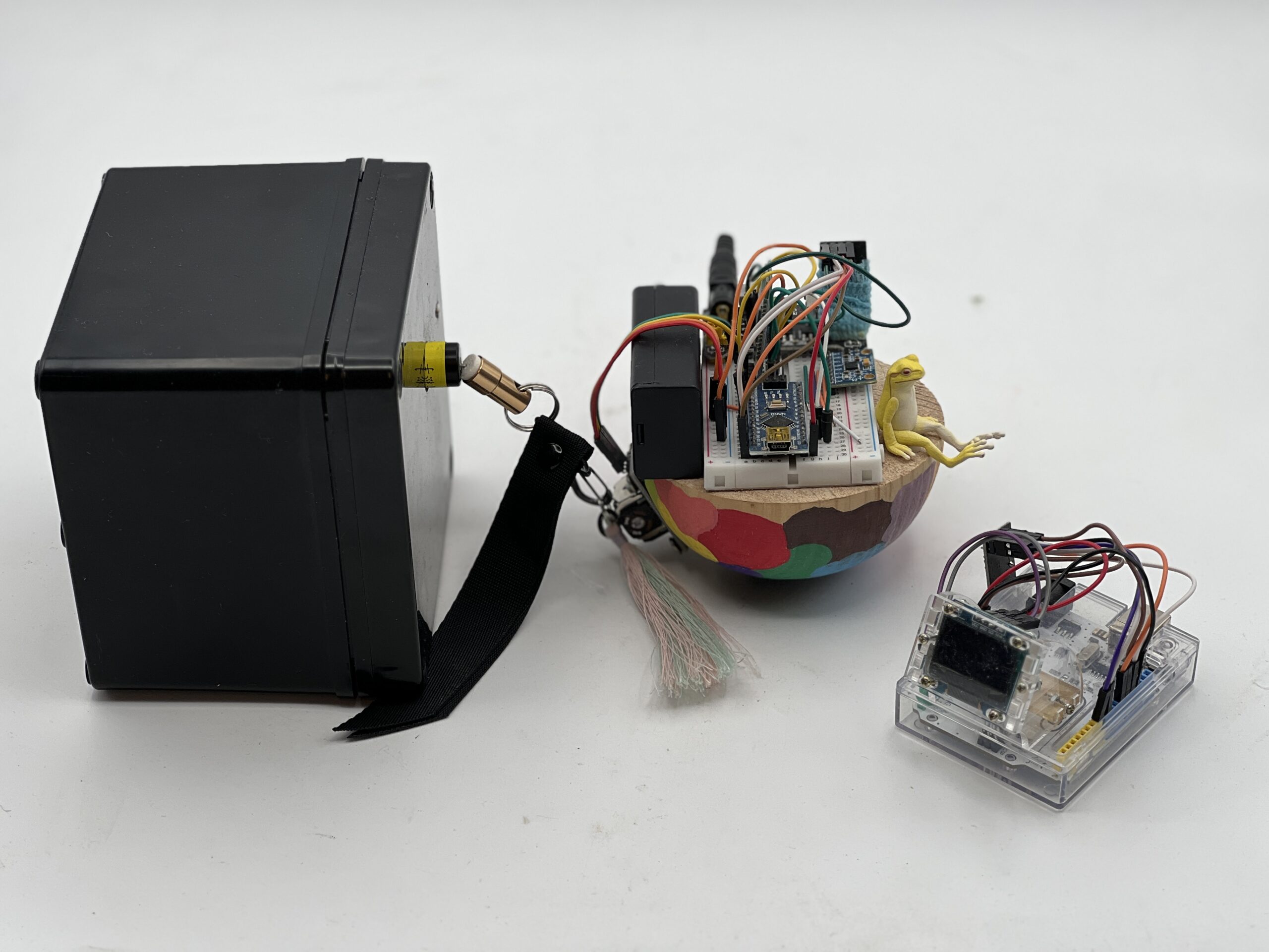 A homemade electronic project with a black battery box, a breadboard with wires and components on a colorful base, and a small clear plastic case containing circuitry, all connected by cables on a white surface.