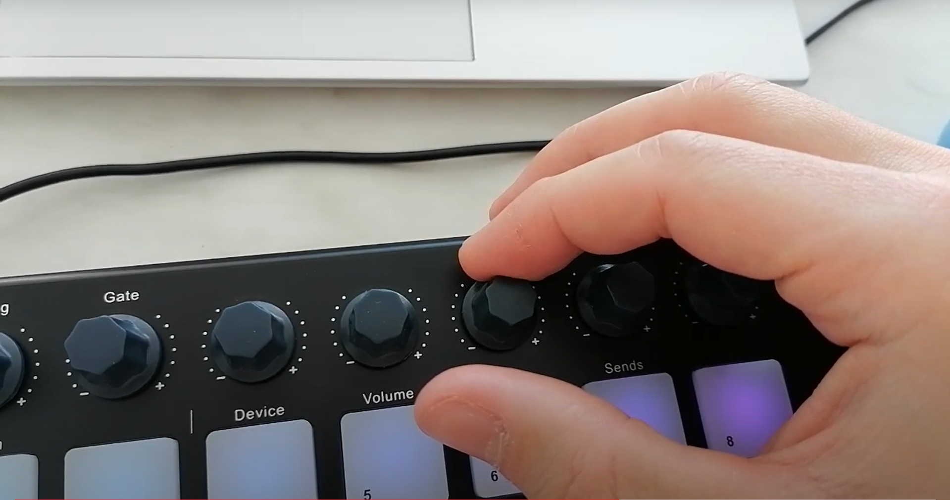 A hand adjusts the volume knob on a black MIDI controller with several other control knobs and illuminated keys visible, in front of a white computer keyboard and a black cable.