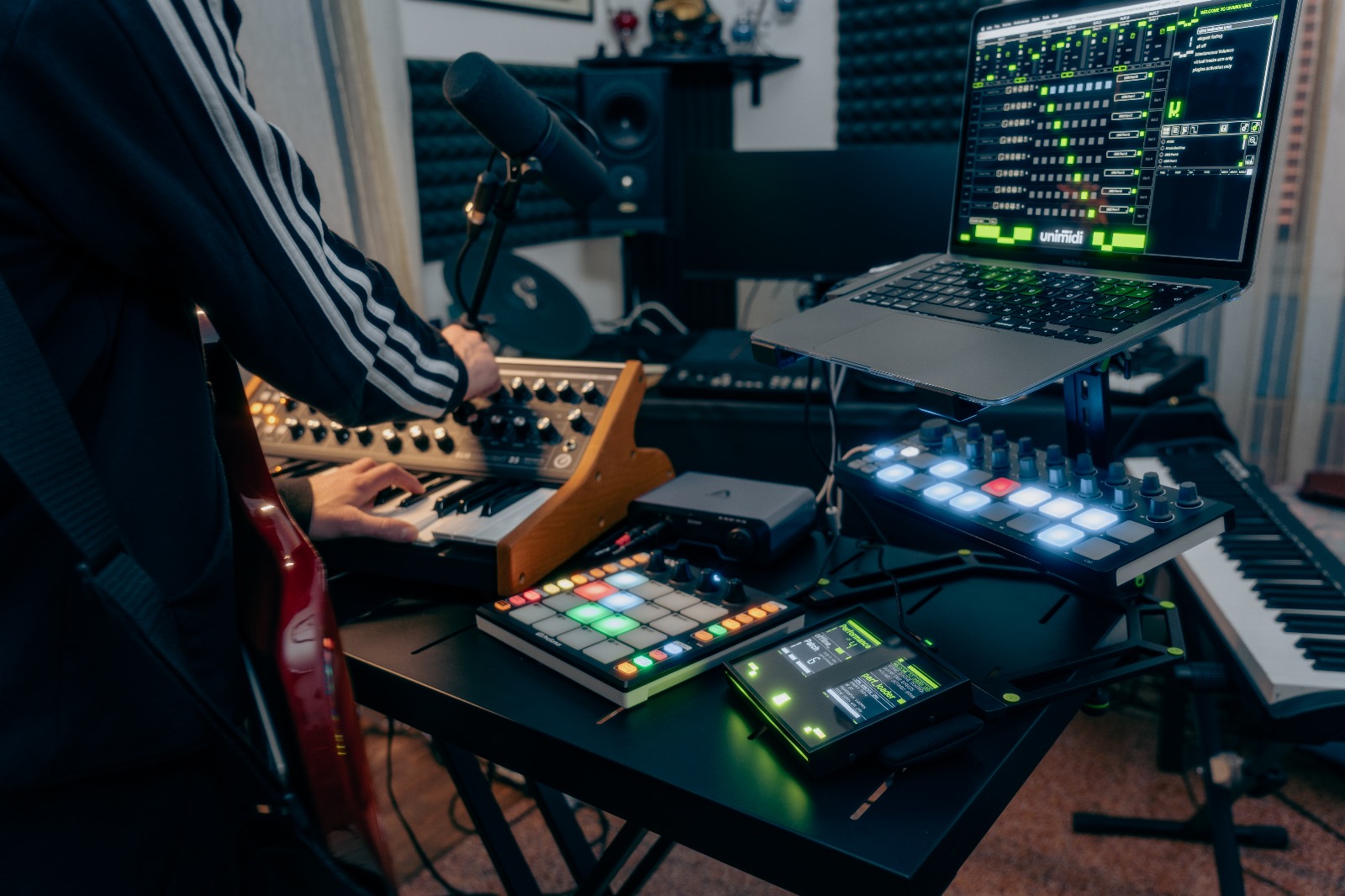 A person operates a synthesizer and music production equipment in a home studio, surrounded by a laptop, MIDI controllers, and various electronic instruments.