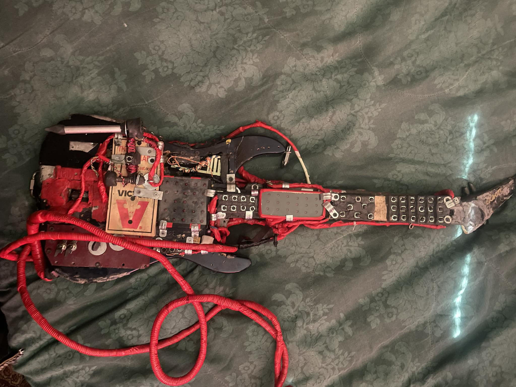 A homemade electric guitar made from mixed electronic components, red wires, circuit boards, and metal plates, lying on a green patterned fabric background.