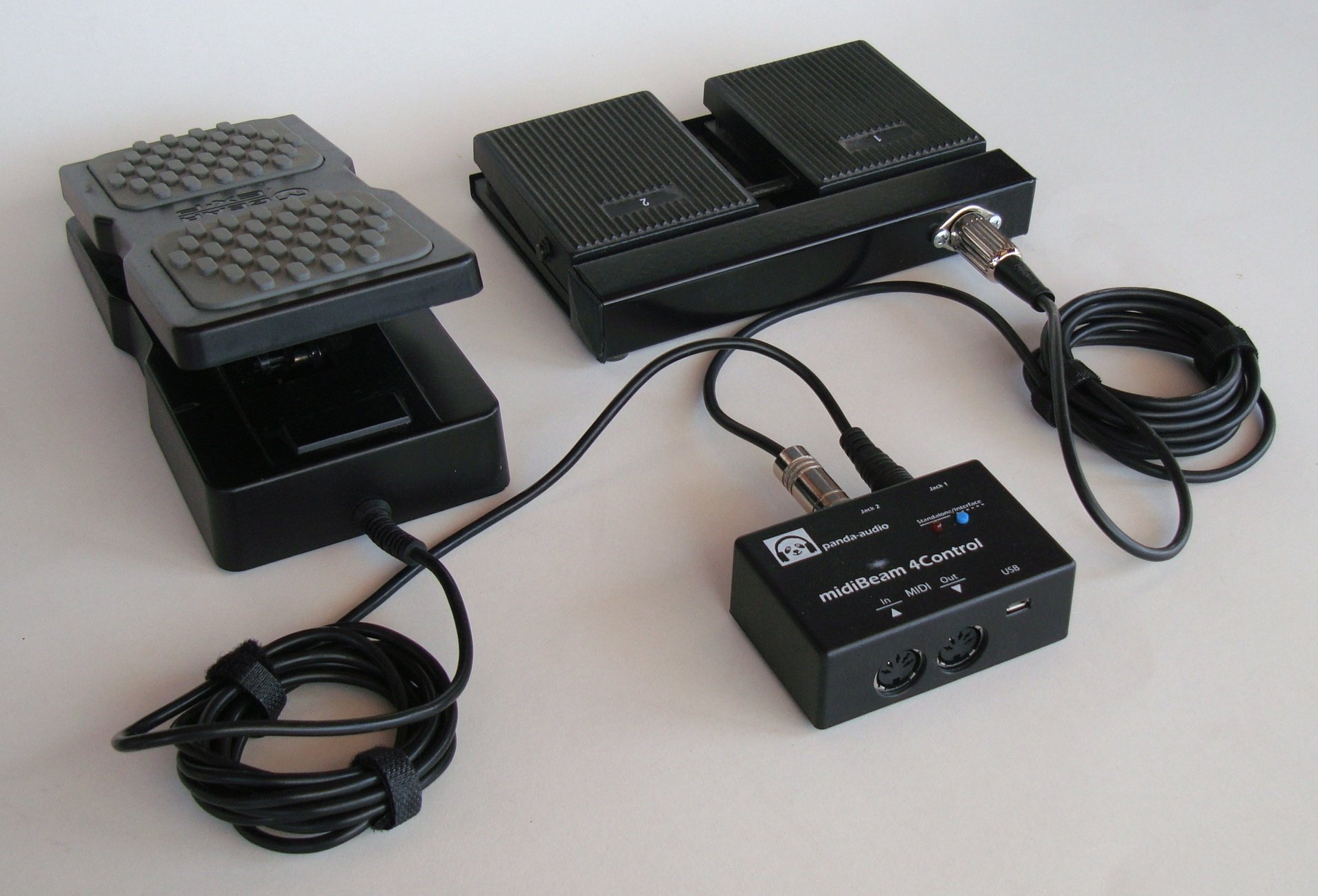 Two black expression pedals are connected to a MIDI controller box with cables. The devices are placed on a light surface, with the cables coiled neatly beside them.