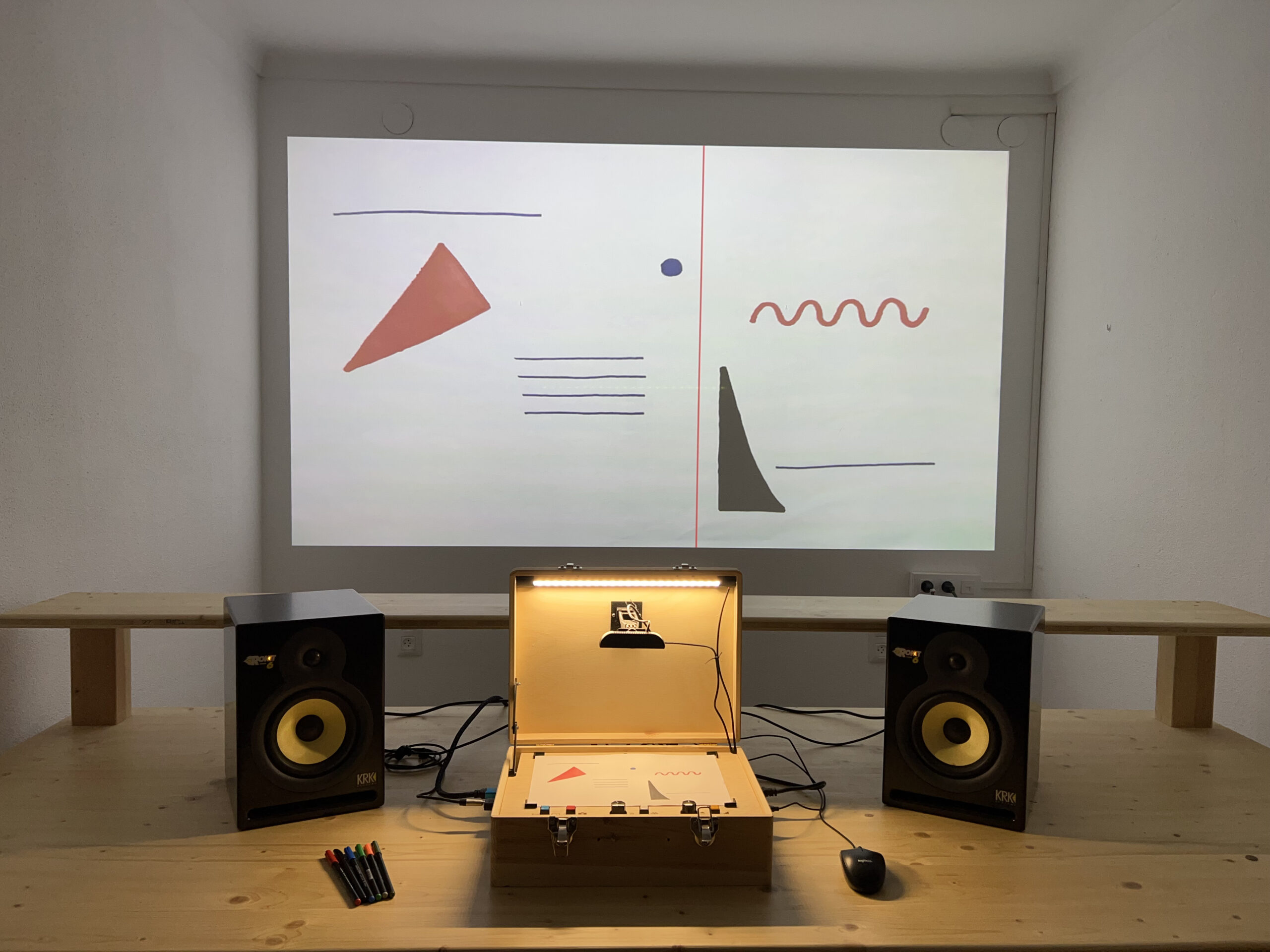 A wooden table holds a briefcase with electronic components, flanked by two speakers. Behind, a wall projection displays simple abstract shapes and lines in red, blue, and black on a white background. Pens and a mouse are on the table.