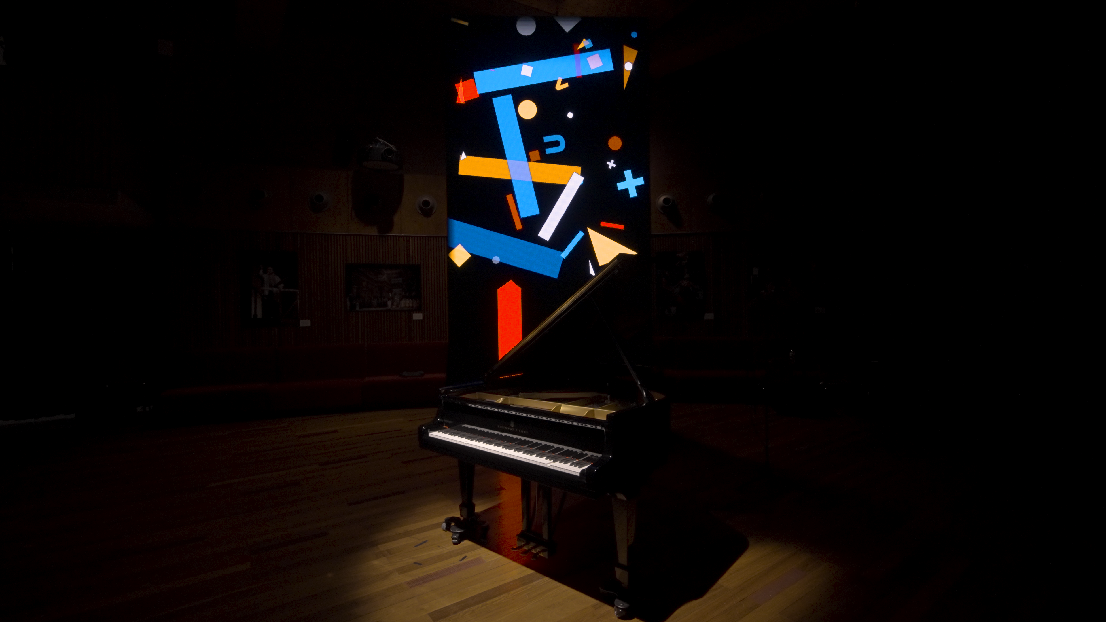 A grand piano sits in a dark room with a colorful, abstract digital projection of geometric shapes and lines displayed above it, casting light onto the piano and wooden floor.