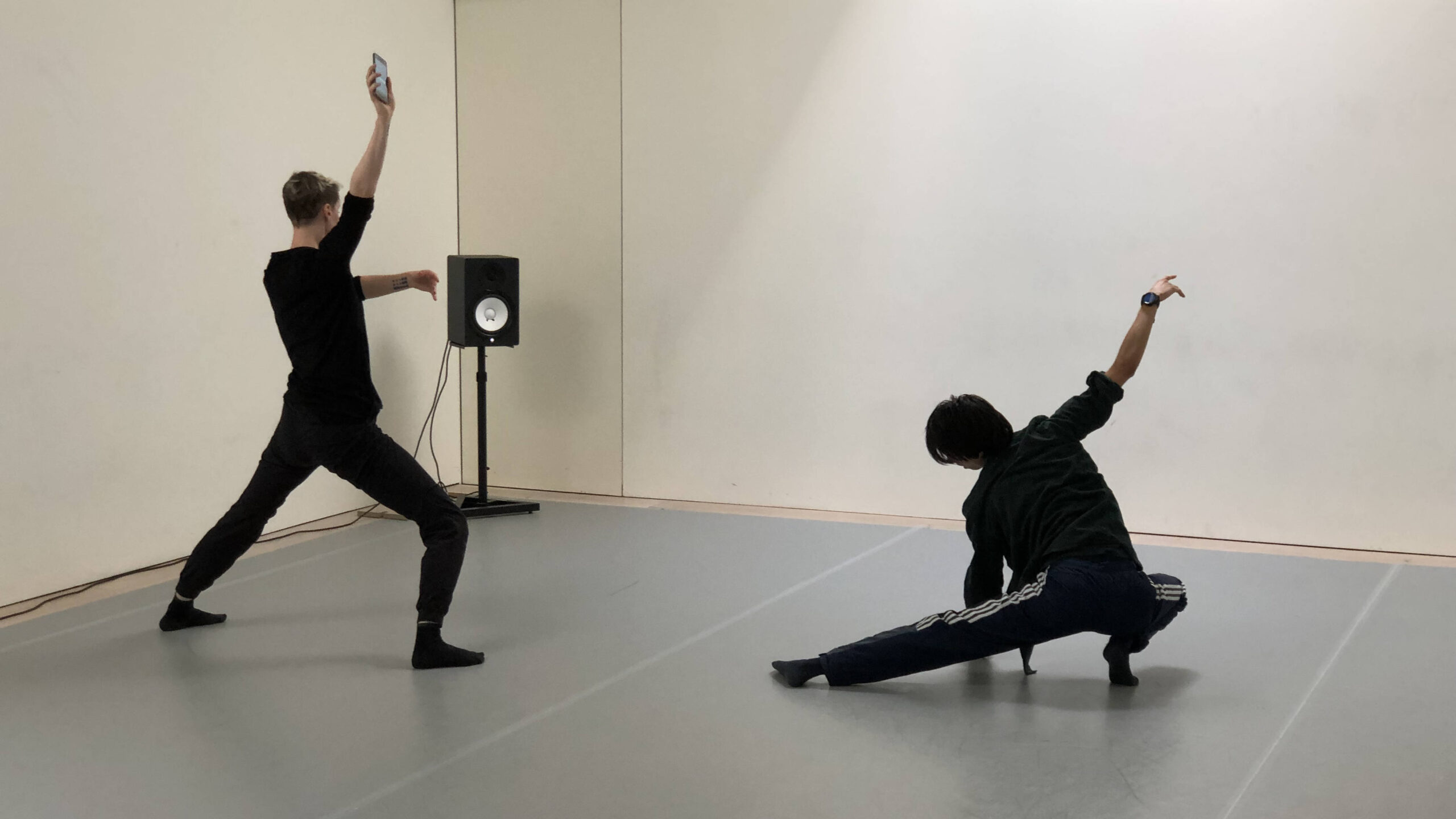 Two people in a dance studio strike dynamic poses, each holding a phone aloft. One stands with legs apart and arm raised, while the other crouches low with an arm extended. A speaker sits against the wall in the background.
