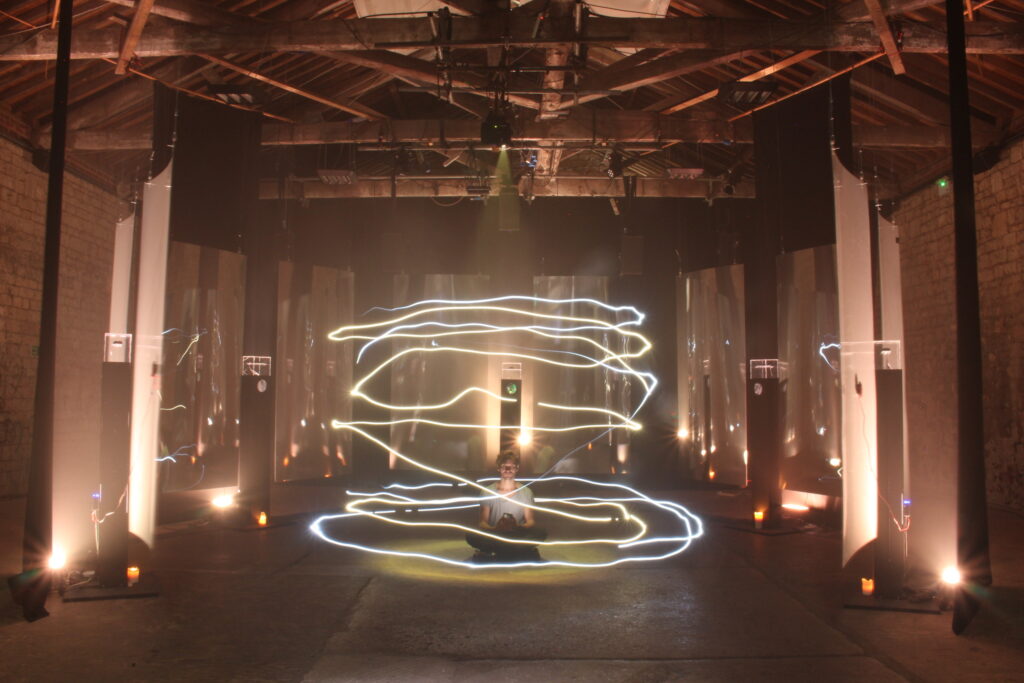 A person sits cross-legged on the floor of a dimly lit industrial space, surrounded by swirling trails of white light and reflective panels, creating an ethereal and immersive atmosphere.