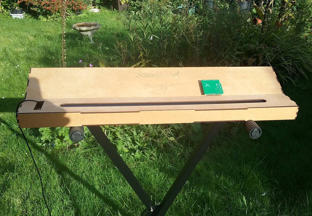 A rectangular wooden device with a slot and a green circuit board sits on a stand outdoors in a sunlit garden with grass and plants in the background.