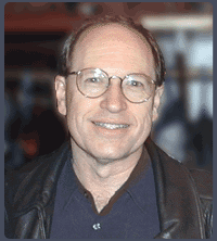 A middle-aged man with glasses and short hair is smiling at the camera. He is wearing a dark shirt and a black leather jacket, with an out-of-focus background behind him.