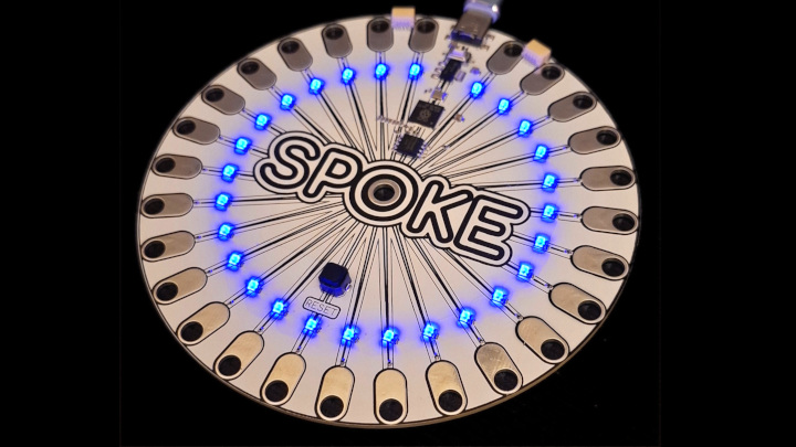 A circular electronic board labeled SPOKE features evenly spaced blue LEDs lit in a ring, USB connector at the top, and various electronic components on a black background.
