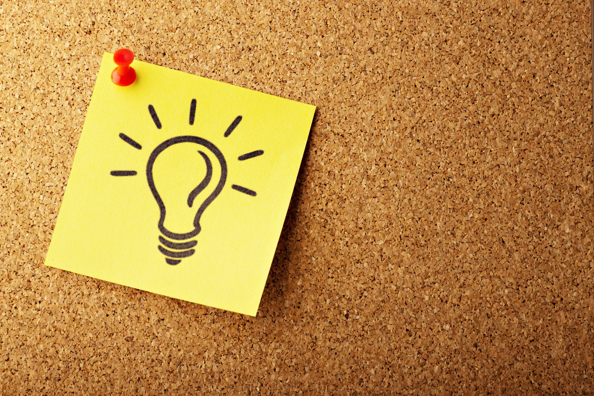 A yellow sticky note with a hand-drawn light bulb and lines representing light is pinned to a corkboard with two red push pins. The note symbolizes an idea or inspiration.