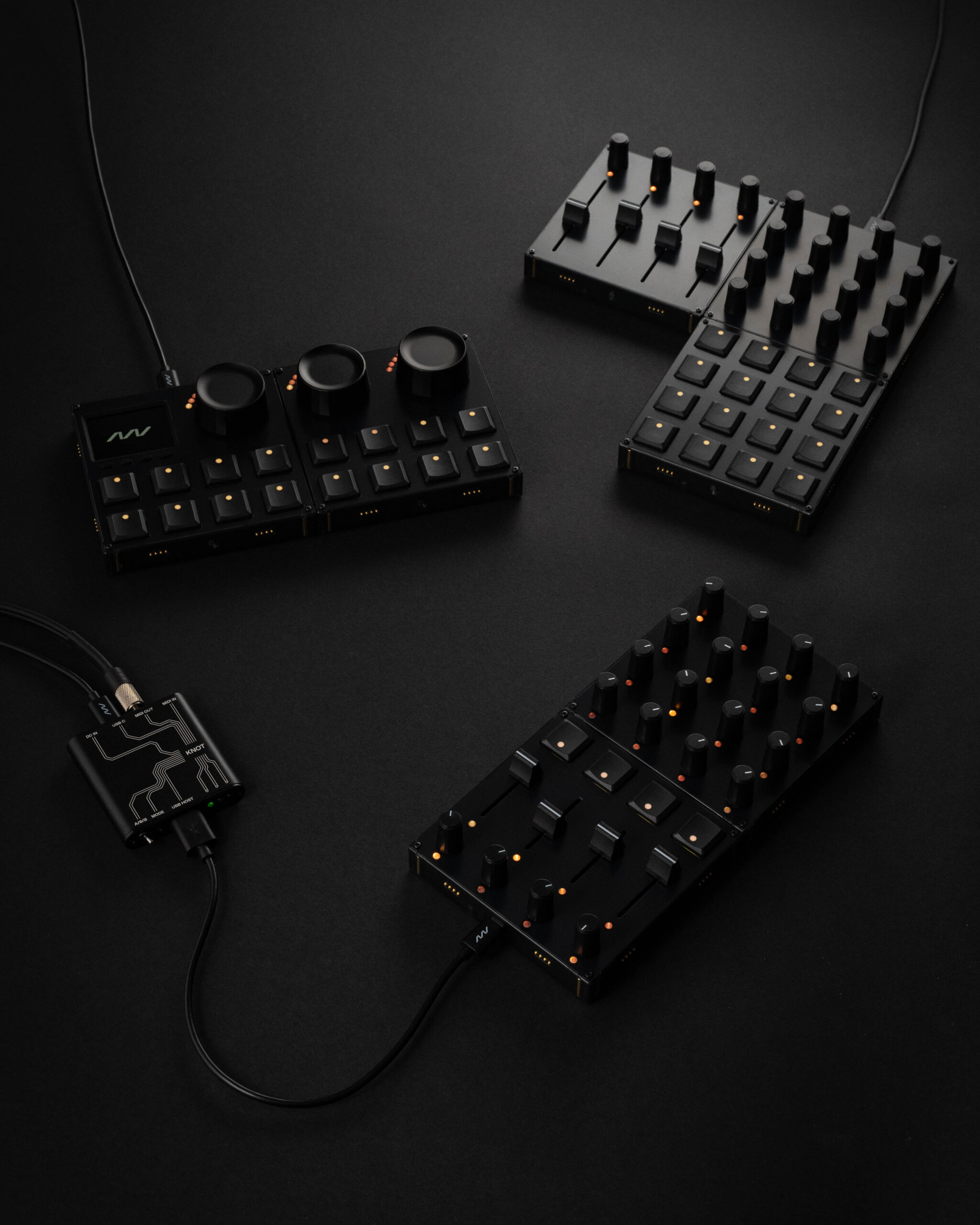 Four modular black MIDI controllers with various knobs, buttons, and dials are arranged on a dark surface, connected by cables, with small orange indicator lights illuminated on each device.