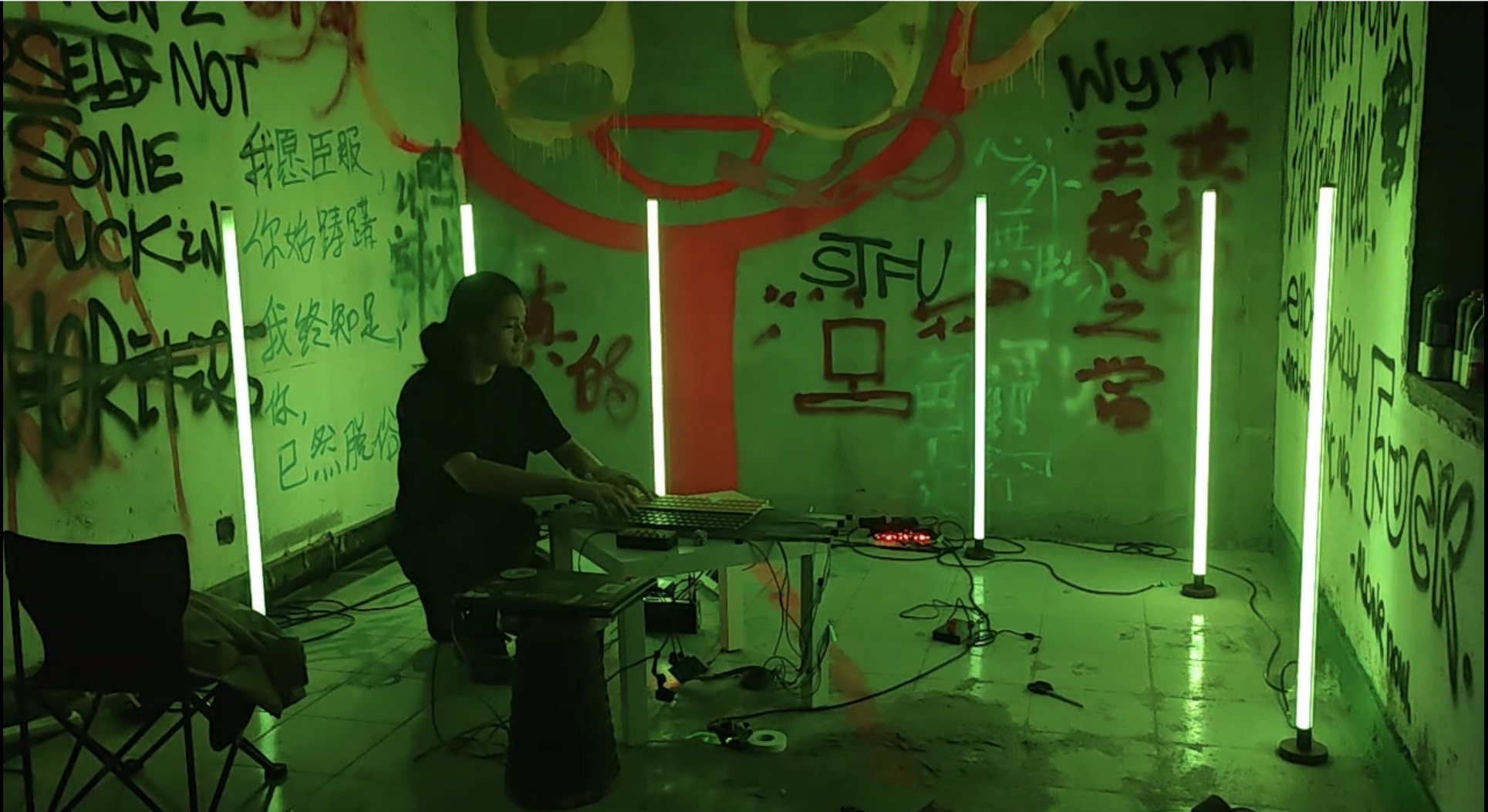 A person sits at an electronic setup in a dark room with bright vertical green lights. The walls are covered in colorful graffiti and Chinese characters. Cables and equipment are scattered across the floor.