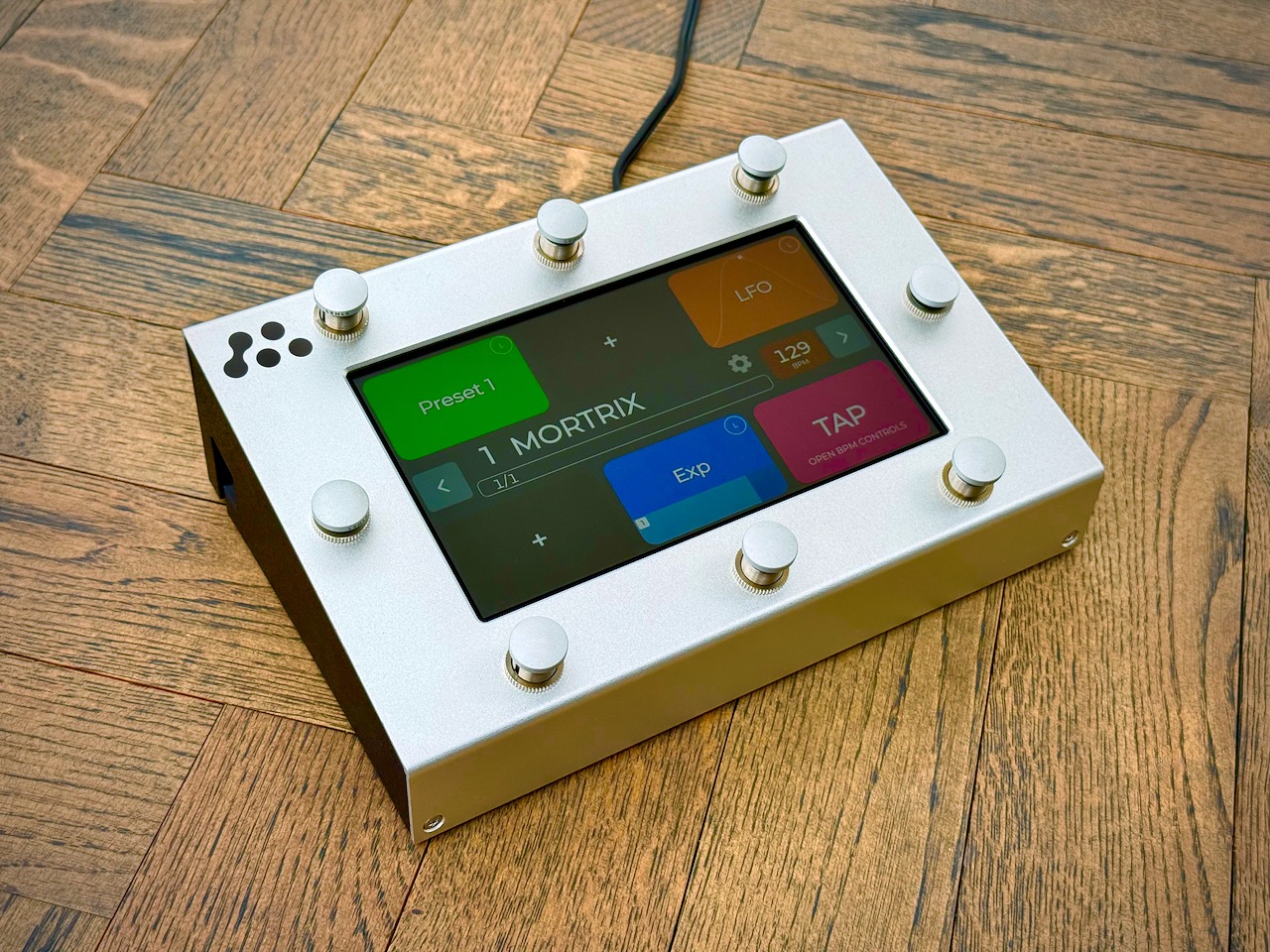 A rectangular electronic device with eight silver knobs and a touchscreen displaying colorful controls sits on a wooden floor. A black cord extends from the back.