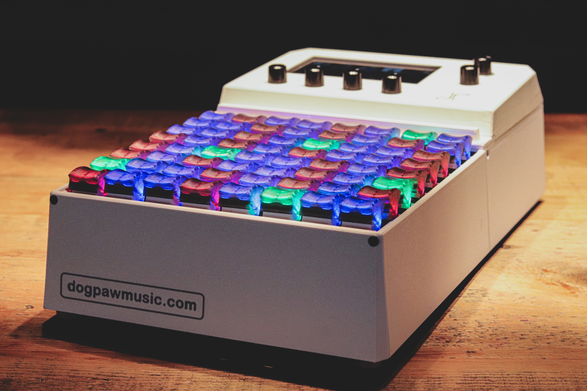 A white electronic music controller with colorful, backlit rubber pads and several black knobs on top, sitting on a wooden surface. A label reads “dogpawmusic.com”.