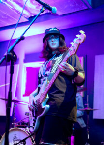A musician wearing a black t-shirt and cap plays an electric bass guitar on stage under purple and blue lights, with a drum set visible in the background.