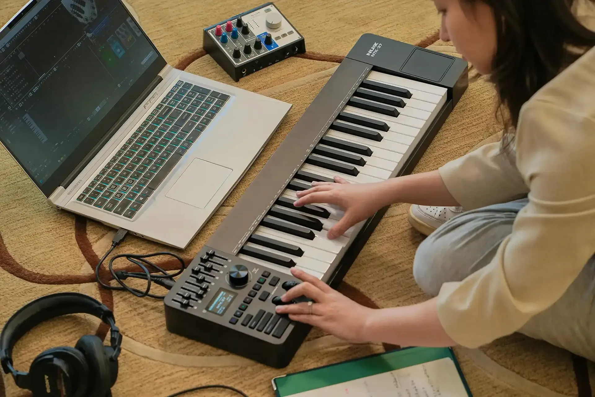 A person sits on the floor playing a digital keyboard next to a laptop, audio interface, and headphones. There is a notebook with handwritten notes nearby, suggesting they are composing or producing music.