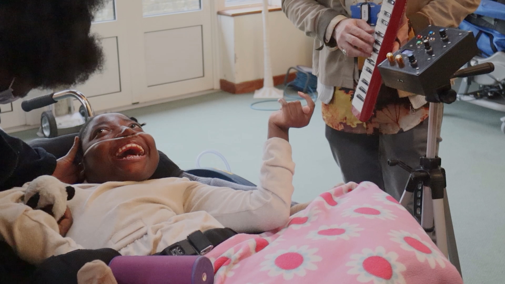 A young person in a wheelchair, covered with a pink daisy blanket, smiles joyfully. Someone beside them plays a keyboard, while another person leans in close, engaging with the child in a bright room.