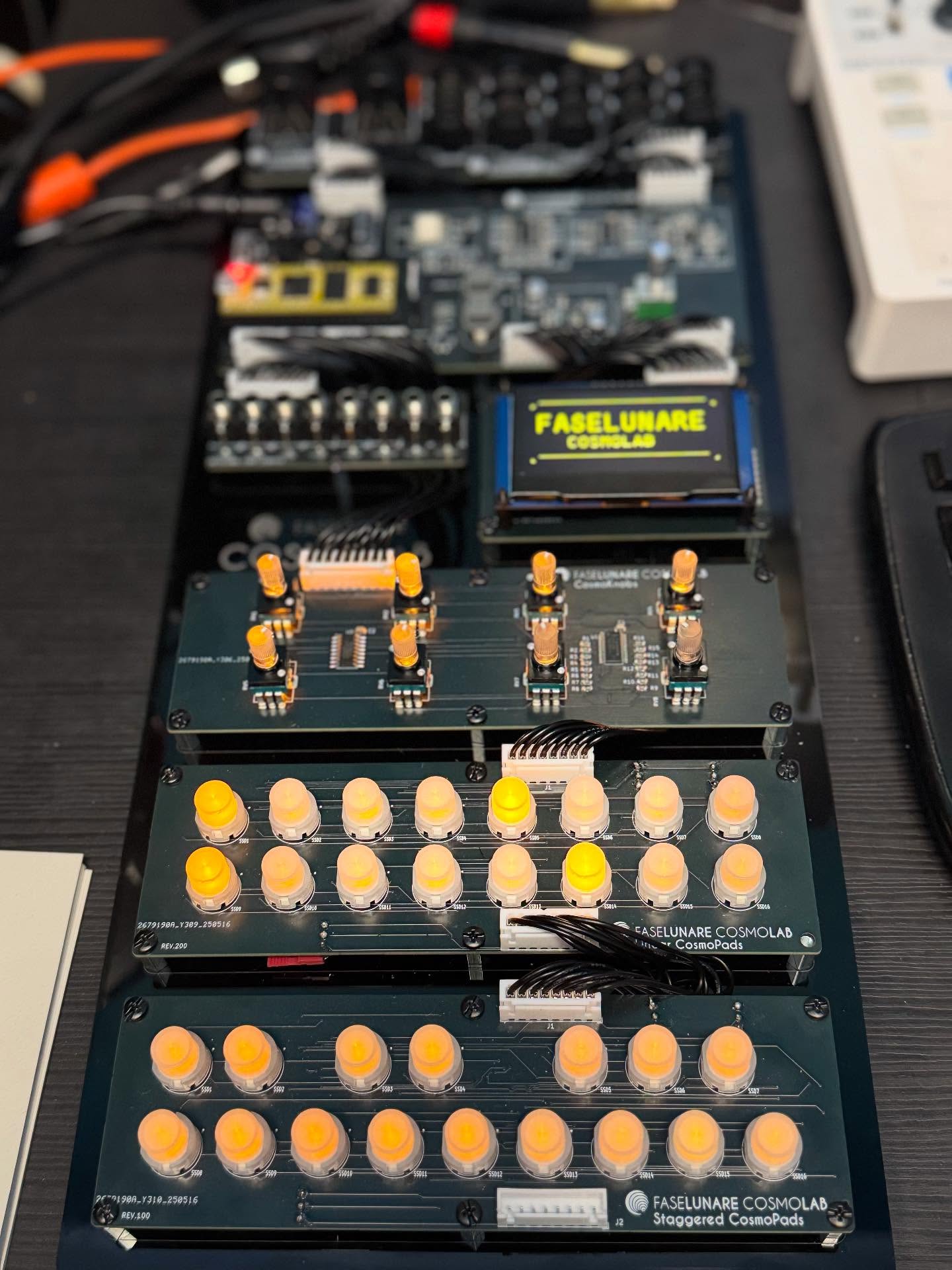A close-up of several modular synthesizer boards with yellow knobs and buttons, electronic components, and patch cables on a dark tabletop. The boards are labeled Faselunare Cosmo.