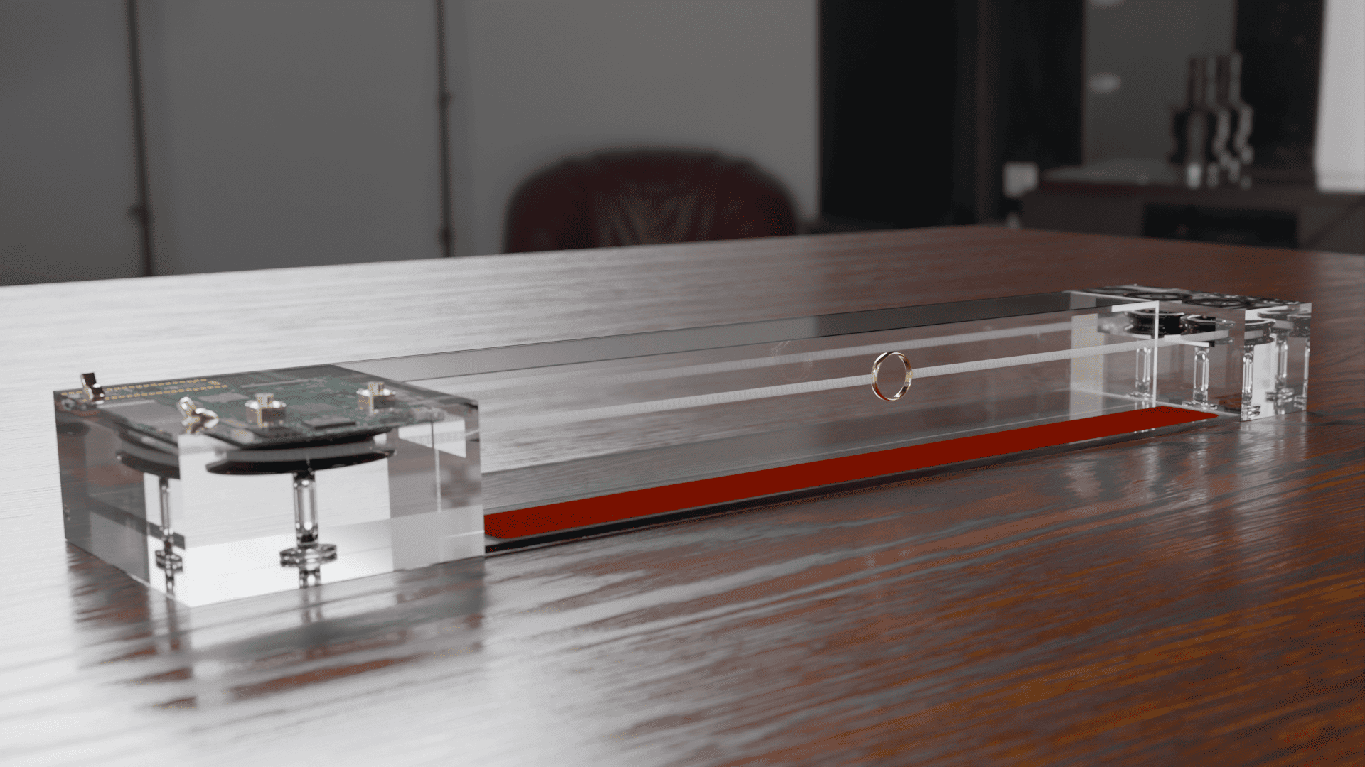A transparent rectangular device with metallic components on each end sits on a wooden table. The device has a clear casing, showing internal mechanisms, and is set in a modern indoor environment.