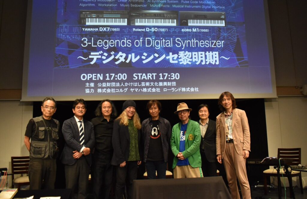 A group of eight people stand on stage in front of a large screen displaying “3-Legends of Digital Synthesizer” with images of synthesizers and event details in English and Japanese; they are smiling and posing for a photo.