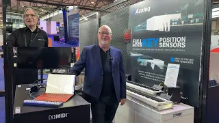 Mike Kent standing at a the Azoteq Music China trade show booth displaying MIDI music equipment and a banner for key position sensors. One man stands by a keyboard and laptop; the other stands behind a counter. Informational posters are visible in the background.