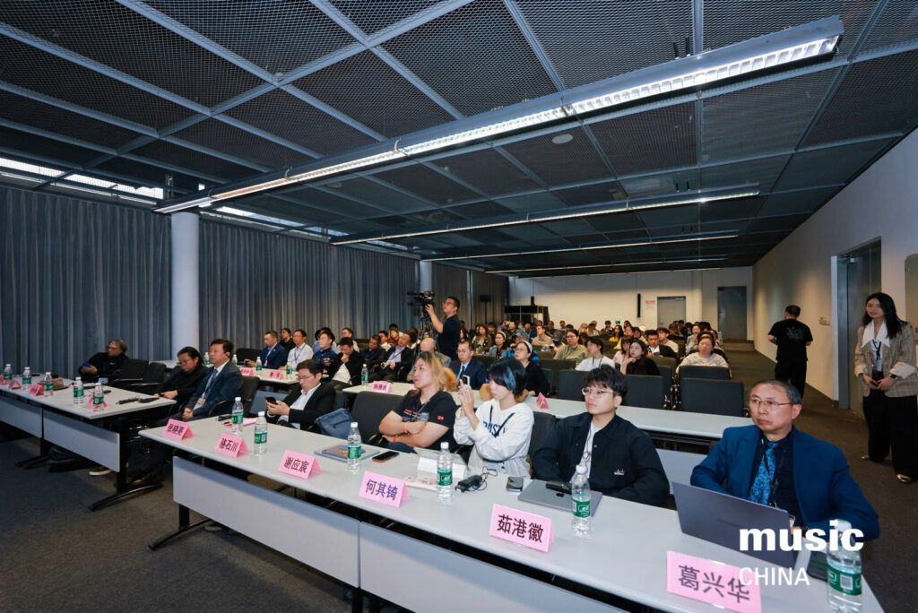 A large group of people sits at conference tables in a modern room, attentively listening to a presentation at Music China. Nameplates, bottled water, and papers are on the tables. The room is brightly lit.